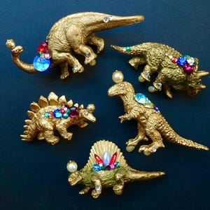 Op de afbeelding: Vijf gouden dinosaurussenfiguren versierd met kleurrijke strass steentjes. De dinosaurussen zijn een stegosaurus, een triceratops, een tyrannosaurus rex, een brachiosaurus en een spinosaurus.