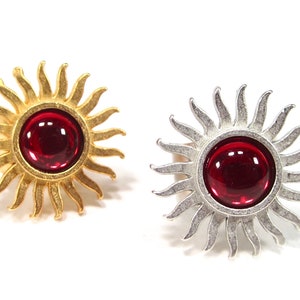 Könnte beinhalten: Zwei sonnenförmige Broschen, eine goldfarbene und eine silberfarbene, jeweils mit einem großen, runden, roten Edelstein in der Mitte. Das Sonnenstrahl-Design hat spitze Strahlen, die sich nach außen erstrecken. Die Broschen werden vor weißem Hintergrund präsentiert.
