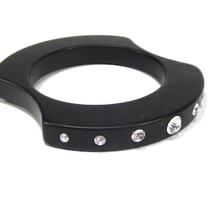 Könnte beinhalten: Ein schwarzes Bangel-Armband mit geschwungenem Design und klaren Strasssteinen.