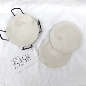 Puede incluir: Tres posavasos redondos de cer&aacute;mica blanca con un dise&ntilde;o de m&aacute;rmol. Los posavasos est&aacute;n apilados uno encima del otro. Los posavasos est&aacute;n hechos a mano por "SASH".