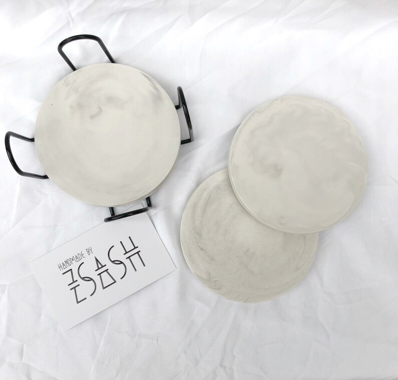 Puede incluir: Tres posavasos redondos de cer&aacute;mica blanca con un dise&ntilde;o de m&aacute;rmol. Los posavasos est&aacute;n apilados uno encima del otro. Los posavasos est&aacute;n hechos a mano por "SASH".