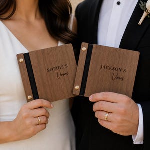 May include: Two wooden vow books with the names "Sophie's" and "Jackson's" engraved on them. The books are held by a couple, showcasing their wedding rings. Each book has a black spine and gold-colored rivets.