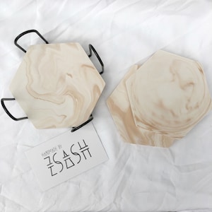 Puede incluir: Dos posavasos hexagonales con un patr&oacute;n de m&aacute;rmol beige. Los posavasos est&aacute;n apilados uno encima del otro. Una etiqueta con el texto "Handmade by ZsaDsh" est&aacute; en la esquina inferior izquierda de la imagen.