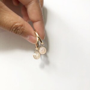 May include: Gold hoop earrings with peach-coloured, faceted gemstones. The earrings are suspended against a white backdrop. The gemstones are small and round.