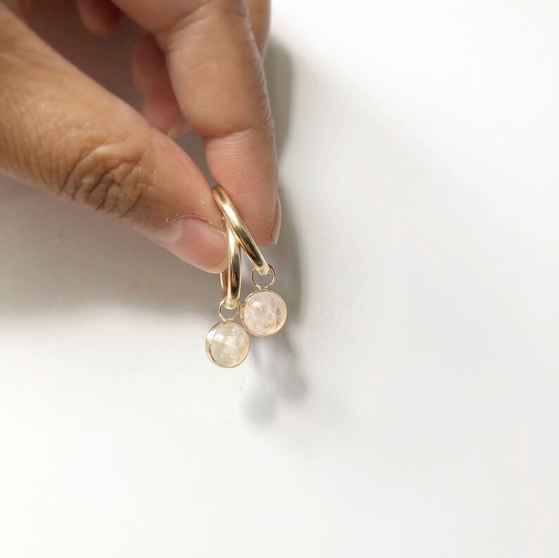 May include: Gold hoop earrings with peach-coloured, faceted gemstones. The earrings are suspended against a white backdrop. The gemstones are small and round.