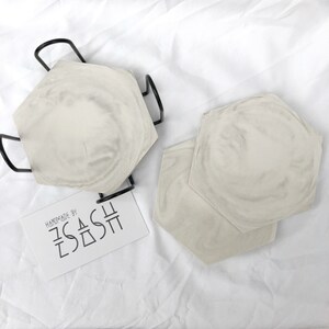 Puede incluir: Conjunto de dos posavasos hexagonales de hormig&oacute;n con un dise&ntilde;o de m&aacute;rmol blanco y gris. Los posavasos est&aacute;n hechos a mano por "ZASH" y se muestran en un soporte de metal negro.