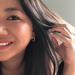 May include: A close-up of a person's face with long, dark brown hair. The person is smiling and wearing a small, gold hoop earring in their left ear.