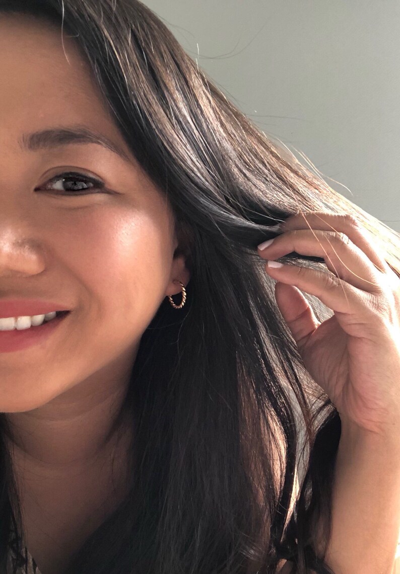 May include: A close-up of a person's face with long, dark brown hair. The person is smiling and wearing a small, gold hoop earring in their left ear.