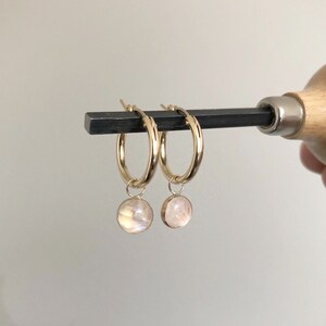 May include: Gold hoop earrings featuring small, round, iridescent moonstone pendants. The earrings are held by a dark metal tool against a neutral background. The hoops are a bright, shiny gold colour.