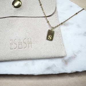 May include: A gold necklace with a small rectangular pendant engraved with the letter "S". The necklace is displayed on a white marble surface and a beige pouch with the text "EACH DASH" embossed on it.