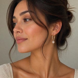 May include: A woman with dark hair in a loose updo, wearing gold hoop earrings with a small, round, white pendant. She also wears a gold necklace and a cream-colored top. The image focuses on her face and jewellery.