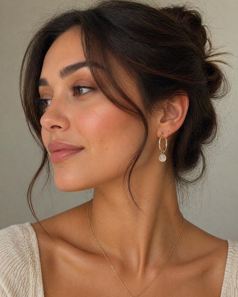 May include: A woman with dark hair in a loose updo, wearing gold hoop earrings with a small, round, white pendant. She also wears a gold necklace and a cream-colored top. The image focuses on her face and jewellery.
