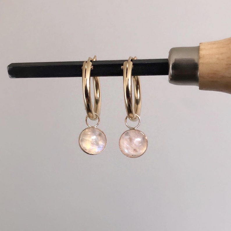 May include: A pair of gold hoop earrings, each with a small, round, iridescent moonstone pendant. The earrings are displayed against a neutral background. The moonstones have a soft, pearly sheen.