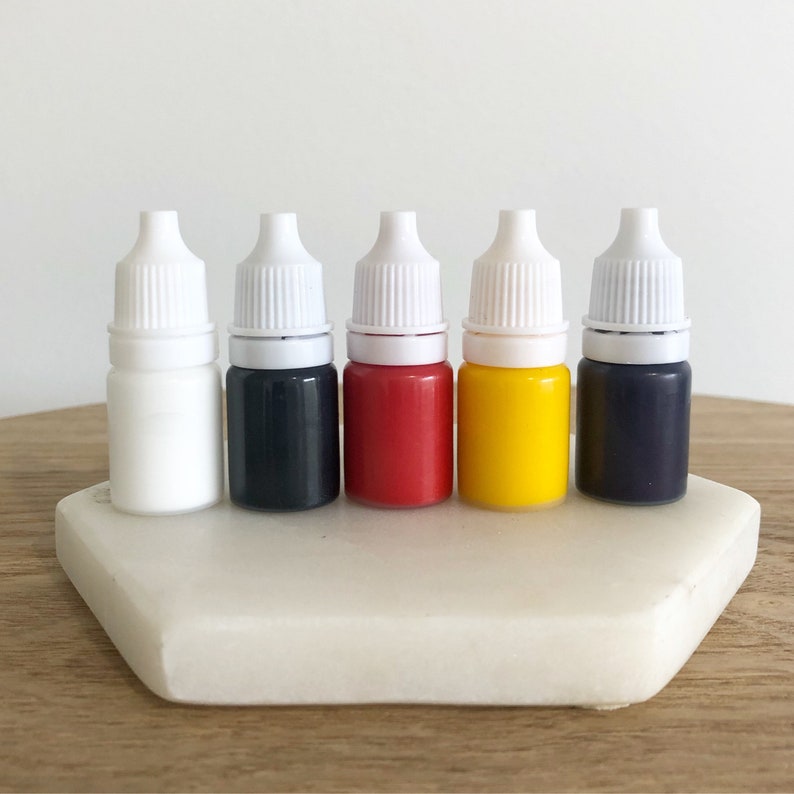 K&ouml;nnte beinhalten: F&uuml;nf kleine Flaschen mit fl&uuml;ssiger Farbe und wei&szlig;en Tropfverschl&uuml;ssen. Die Flaschen sind von links nach rechts angeordnet: wei&szlig;, schwarz, rot, gelb und blau.