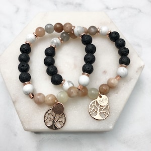 May include: Two bracelets featuring black lava rock beads, white howlite beads, and sunstone beads. Each bracelet has a gold tree of life charm with the word "JASH" engraved on one charm.