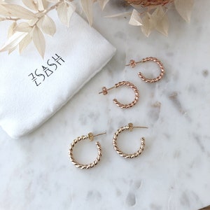 May include: Two pairs of gold hoop earrings with a twisted design. The earrings are on a white marble surface. The earrings are packaged in a white pouch with the text "The Dash" printed on it.