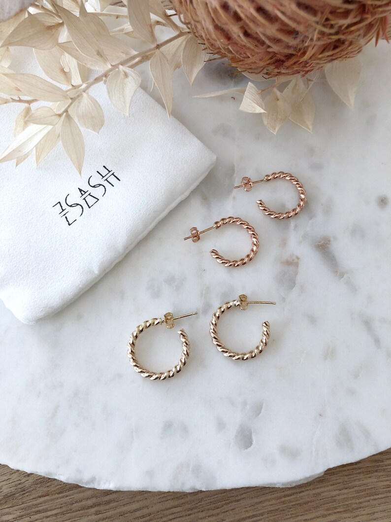 May include: Two pairs of gold hoop earrings with a twisted design. The earrings are on a white marble surface. The earrings are packaged in a white pouch with the text "The Dash" printed on it.