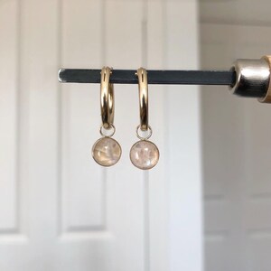 May include: Two gold hoop earrings, each with a small, round, iridescent stone pendant. The earrings are displayed on a dark metal bar. The stones have a soft, pearly glow, and the hoops are a bright gold colour.