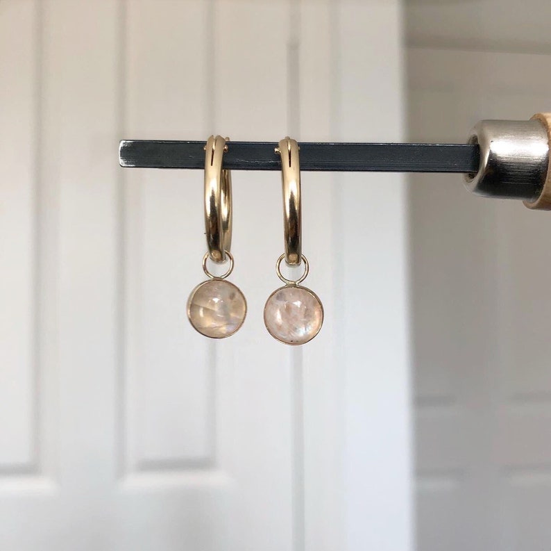 May include: Two gold hoop earrings, each with a small, round, iridescent stone pendant. The earrings are displayed on a dark metal bar. The stones have a soft, pearly glow, and the hoops are a bright gold colour.