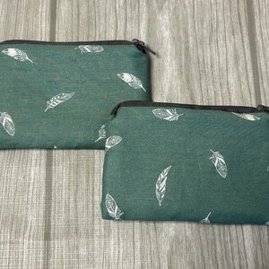 Peut inclure: Deux pochettes zippées de couleur sarcelle avec un motif de plumes blanches. Les pochettes sont rectangulaires, avec des fermetures éclair noires et un design simple. Elles sont probablement utilisées pour ranger de petits objets.