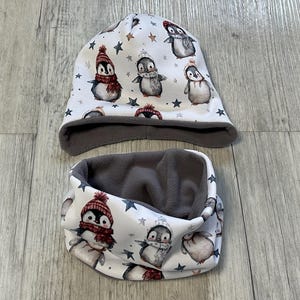 May include: A white beanie hat and neck warmer set featuring a penguin design. The penguins are depicted wearing hats and scarves, with a grey fleece lining. The pattern includes stars, ideal for colder weather.