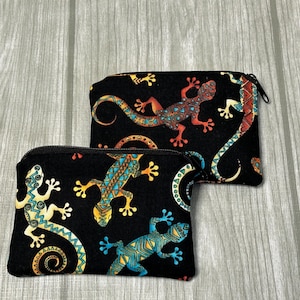 May include: Two black zippered pouches with a colorful lizard pattern. The lizards are in shades of red, orange, yellow, blue, and green. The pouches are rectangular and appear to be made of fabric.