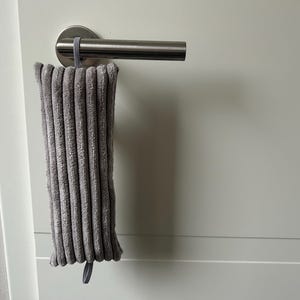 May include: A grey, corduroy door handle cover hangs from a silver door handle. The cover is rectangular with vertical ridges and a small loop at the bottom. The door and door frame are white.