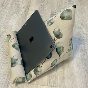 Könnte beinhalten: Ein graues Tablet liegt in einem beigen Kissenständer. Das Kissen ist mit einem grünen und grauen Blättermuster verziert. Der Ständer ist so konzipiert, dass er ein Tablet in einem Winkel für freihändiges Betrachten hält.