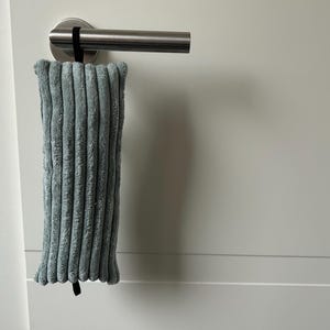 May include: A pale blue corduroy door silencer hangs from a silver door handle. The rectangular silencer has a ribbed texture and is vertically oriented. A black strap secures the silencer to the handle. The door and wall are white.