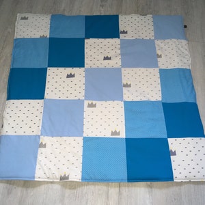 Könnte beinhalten: Ein blau-weißes Patchwork-Quilt mit einem quadratischen Muster. Die Quadrate sind entweder einfarbig blau, gepunktet oder mit Kronen verziert. Diese Decke, wahrscheinlich für ein Baby oder Kind, ist aus weichem Stoff.