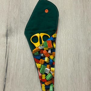 May include: A green fabric scissor holder with a colourful pattern of toy building blocks. The holder is open, revealing yellow scissors inside. The holder has a snap closure and is on a light wood surface.