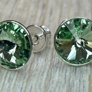 Op de afbeelding: Close-up van twee ronde, lichtgroene kristallen oorstekers met een facetgeslepen design. De oorbellen hebben een zilverkleurige metalen zetting en pin. De achtergrond is een lichte houtnerf.