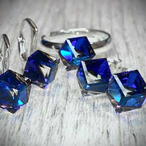 Pode incluir: Um conjunto de joias com um anel e brincos com detalhes de cristal azul profundo facetado. Os brincos são do estilo de alavanca. O anel e os brincos têm uma moldura de metal prateado. Os cristais têm uma forma geométrica, semelhante a um cubo.