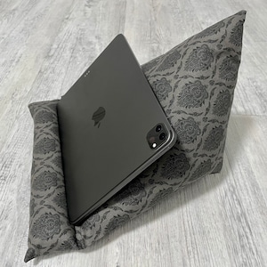 Könnte beinhalten: Ein graues Tablet liegt in einem grau gemusterten Kissenständer. Das Kissen hat ein sich wiederholendes Blumenmuster. Das Tablet ist dunkelgrau mit einer schwarzen Kamera und dem Apple-Logo.