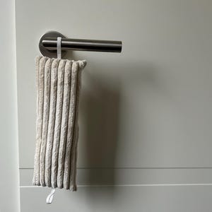 May include: A beige, corduroy-textured rectangular item hangs from a silver door handle. The item is suspended by a white loop. The background is a light grey wall and door frame.