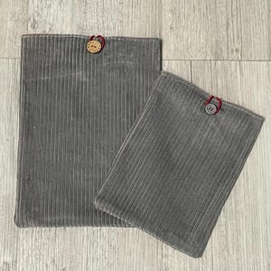 Könnte beinhalten: Zwei graue Cord-Beutel in verschiedenen Größen. Jeder Beutel hat einen Knopfverschluss mit roter Schlaufe. Der größere Beutel ist rechteckig, der kleinere quadratisch. Der Cord hat vertikale Rippen.
