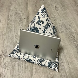 Könnte beinhalten: Ein graues iPad liegt auf einem weißen und marineblauen Kissenständer mit Gaming-Motiv. Der Ständer hat ein sich wiederholendes Muster aus Arcade-Spielgrafiken und Gamer-Gesichtern. Das Kissen ist für die freihändige Nutzung eines Tablets konzipiert.