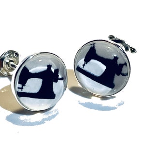 Könnte beinhalten: Ein Paar silberfarbene Ohrstecker. Jeder Ohrring hat einen klaren, runden Glaskabochon mit einer schwarzen Silhouette einer alten Nähmaschine. Die Ohrringe sind vor einem hellweißen Hintergrund platziert.