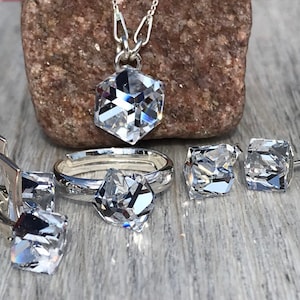 Pode incluir: Um conjunto de joias de prata com pedras de cristal transparente facetadas. O conjunto inclui um colar com pingente, um anel e dois pares de brincos. Os brincos são de estilo alavanca e tachas. O anel e o pingente têm formato hexagonal.