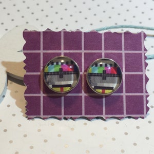 Op de afbeelding: Een paar zilverkleurige oorstekers. Elke oorbel heeft een ronde, heldere glazen cabochon met een kleurrijk tv-testpatroonontwerp. De oorbellen worden gepresenteerd op een paars papier met een rasterpatroon.