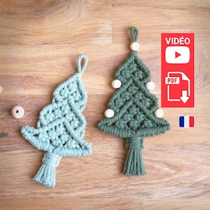Peut inclure: Deux décorations de Noël en macramé en forme d'arbre, l'une verte et l'autre bleu clair, avec des perles en bois blanches et des pompons. Les décorations sont sur une surface en bois. L'image comprend également une boîte rouge avec le texte "VIDÉO" et un bouton de lecture, une boîte blanche avec le texte "PDF" et une flèche de téléchargement, et un drapeau français.