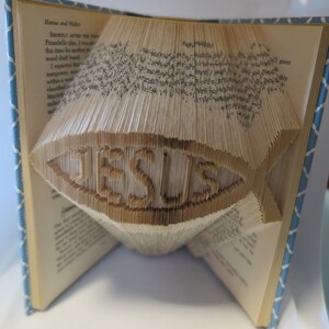 May include: A book folded into the shape of a fish with the word "JESUS" inscribed in the center. The book is open and the pages are folded to create the fish shape.