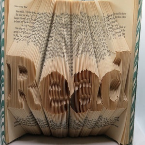 May include: A book art piece with the word "Read" folded into the pages. The book is open and the pages are folded to create the letters. The book is on a white surface.