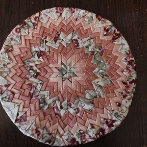 May include: A round, pink and green fabric coaster with a starburst pattern. The coaster is made of many small pieces of fabric sewn together.