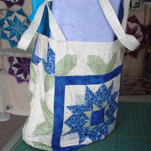 Peut inclure: Un sac fourre-tout en tissu blanc avec des motifs de patchwork vert et bleu. Le sac a un motif d'étoile bleue avec un centre blanc et une bordure bleue. Le sac est posé sur un tapis de coupe vert.