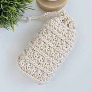May include: A cream-coloured, crocheted soap saver bag with a drawstring closure. The bag features a textured, star-like pattern and a loop for hanging. A bar of soap is partially visible in the background, along with a small green plant.