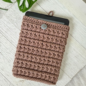 May include: A handmade, crochet tablet sleeve in a muted mauve color. The sleeve features a textured stitch pattern and a button closure. The top has a small loop for easy removal. The sleeve is designed to protect a tablet or e-reader.