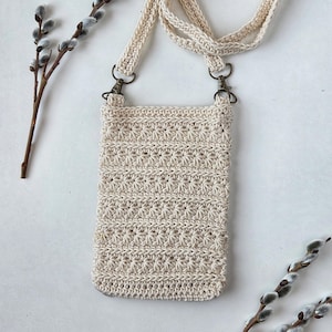 May include: A cream-coloured, crocheted crossbody phone bag with a textured starburst pattern. The bag has a long, braided strap and metal clasps. The bag is rectangular and appears to be handmade.