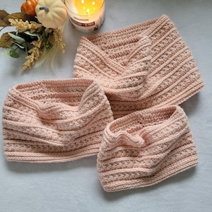 May include: Three handmade, pale pink crochet cowls in different sizes. The cowls have a textured stitch pattern with horizontal stripes and star-like details. A lit candle and small pumpkins are in the background.