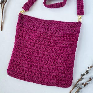 May include: A handmade, magenta crochet crossbody bag with a textured stitch pattern. The bag features a long, adjustable strap and gold-colored metal hardware. The bag is approximately 10 inches wide and 12 inches tall.